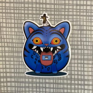 Peut inclure: Autocollant de chat bleu avec des yeux orange, des dents pointues et une langue tirée. Le chat a des rayures noires et un petit oiseau portant un chapeau sur la tête. La langue du chat a un petit écran avec une image de chat.