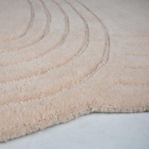 May include: A close-up of a beige rug with a curved, textured design. The rug has a soft, plush appearance and a neutral colour palette, suitable for various interior styles. The design features multiple parallel curved lines.