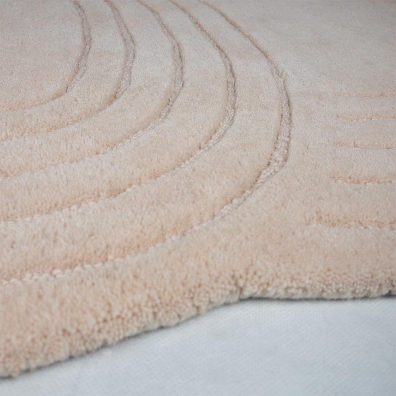 May include: A close-up of a beige rug with a curved, textured design. The rug has a soft, plush appearance and a neutral colour palette, suitable for various interior styles. The design features multiple parallel curved lines.