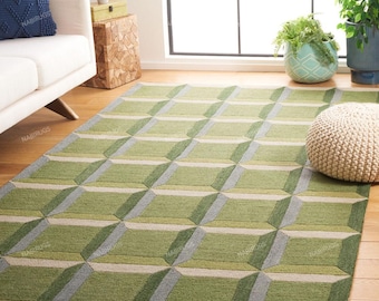 Puede incluir: Una alfombra con estampado geométrico verde y gris. La alfombra presenta un diseño de cubo 3D repetido en tonos verdes, grises y beige. La alfombra está colocada sobre un suelo de madera, con un sofá blanco y plantas al fondo.