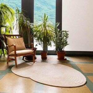 May include: A cream oval rug with a concentric circle pattern on a tiled floor. A wooden armchair with a beige cushion is to the left, and potted plants are in the background. The rug adds a soft texture to the room.