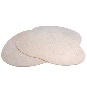 May include: Three beige oval rugs with concentric, curved lines. The rugs are arranged in a layered pattern, with the top rug partially overlapping the others. The rugs have a soft, plush texture and a neutral colour palette.