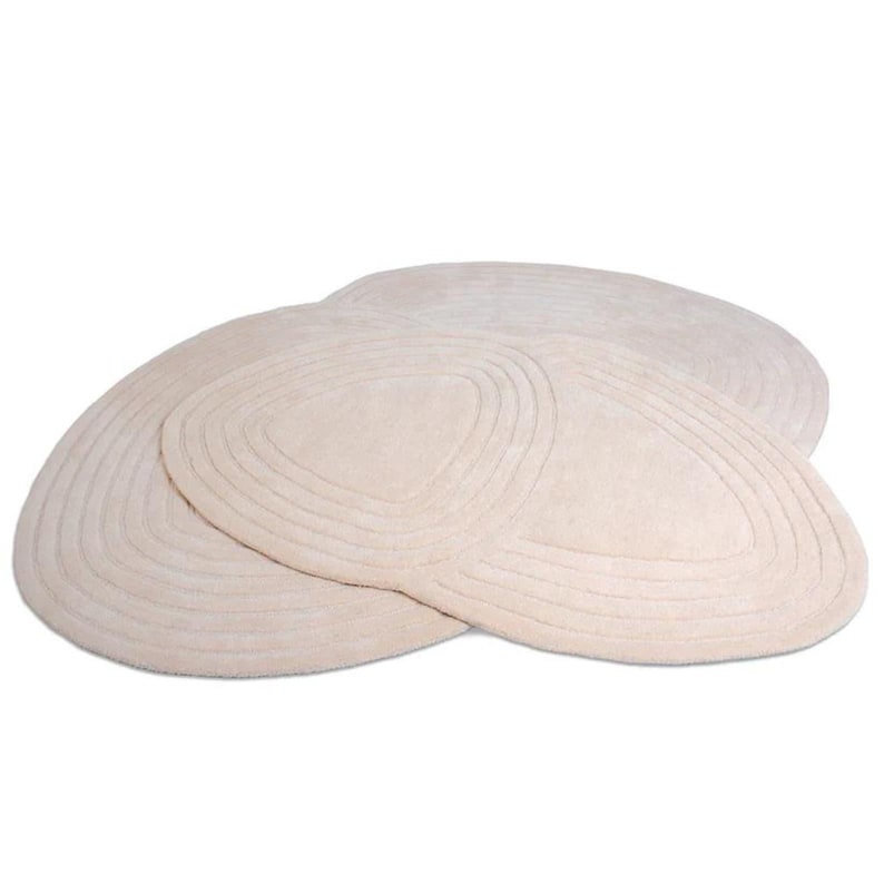 May include: Three beige oval rugs with concentric, curved lines. The rugs are arranged in a layered pattern, with the top rug partially overlapping the others. The rugs have a soft, plush texture and a neutral colour palette.