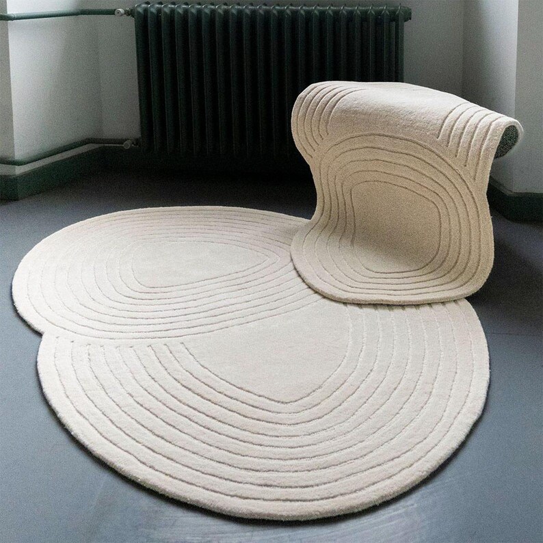 May include: A set of cream-coloured rugs, comprising a large circular rug and a smaller, uniquely shaped rug. Both rugs have a textured, ridged design with concentric lines. The rugs are on a grey floor, with a dark green radiator in the background.