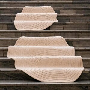 May include: Beige stair treads with a textured, wave-like pattern. The treads are placed on stone steps, creating a soft contrast with the hard surface. The design adds a touch of warmth and style to the staircase.