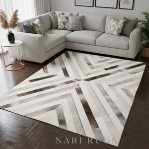 May include: A large, geometric patterned rug with a mix of white, gray, and brown tones. The rug is placed in front of a light gray sectional sofa. The text "NABI RUGS" is visible on the rug.