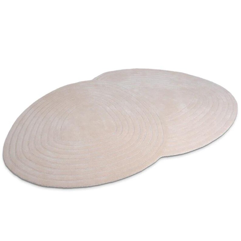 May include: A light beige, oval rug featuring a concentric, ridged pattern. The rug is made from a soft, plush material. The neutral colour of the rug would complement a variety of interior design styles, adding warmth to a room.