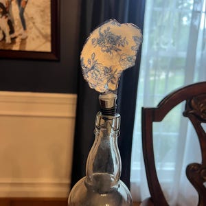 May include: A clear glass bottle with a decorative bottle stopper. The stopper has a cream-coloured surface with a blue floral design. The bottle is partially filled and sits on a table.
