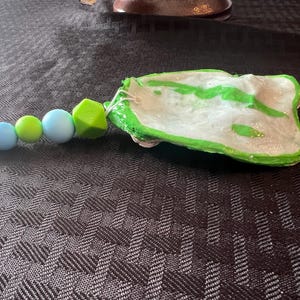May include: A handmade, white and green painted, seashell-shaped item with a beaded strand. The strand features green, blue, and light blue beads, along with a green geometric bead. The item is on a dark, textured surface.
