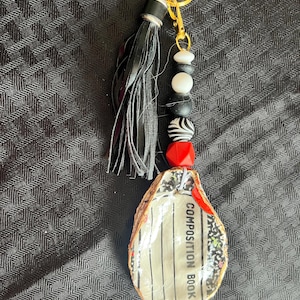 May include: A handmade keychain with a black leather tassel, a gold-coloured clasp, and beads in black, white, and red. The keychain has a pendant shaped like a composition book with the words "COMPOSITION BOOK".