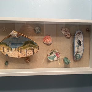 May include: A white framed shadow box displaying various seashells and painted clam shells. One shell is painted with a beach scene, and another depicts a bird. The shells are arranged on a beige fabric background.