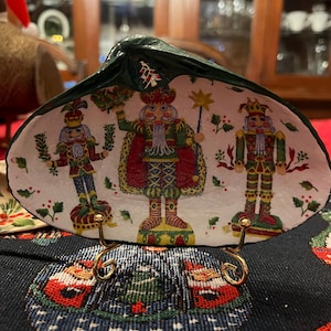Puede incluir: Un plato de concha pintado a mano con tres figuras de cascanueces con atuendos festivos. El interior de la concha es blanco con detalles rojos, verdes y dorados. El plato está sostenido por un soporte de metal dorado, sobre un mantel navideño.