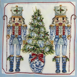May include: A decorative napkin featuring two blue and white nutcracker soldiers flanking a decorated Christmas tree. The tree is in a blue and white vase with a red bow. The napkin has a red border.