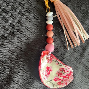 May include: A handcrafted keyring with a gold-tone clasp and a pink tassel. The keyring features a beaded strand with various coloured beads and a painted oyster shell pendant with red floral designs.