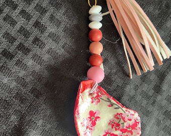 May include: A handmade keychain with a gold-colored clasp and a pink tassel. The keychain features a beaded strand with various colored beads and a painted oyster shell pendant with red floral designs.