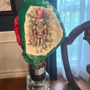 Può includere: Un tappo decorativo per bottiglia con un'immagine dipinta di un soldato schiaccianoci. Il tappo ha un bordo rosso e verde ed è attaccato a una bottiglia di vetro trasparente. Lo schiaccianoci indossa un'uniforme e un cappello rossi.