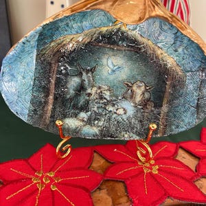 Può includere: Una conchiglia decorativa con una scena della natività dipinta. Il guscio è sostenuto da due supporti in metallo color oro. Decorazioni di poinsettia rosse con accenti dorati in primo piano. Il guscio ha un bordo dorato.