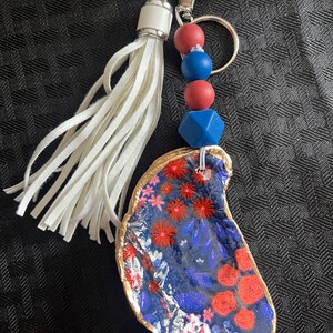 May include: A handcrafted keychain with a painted oyster shell. The shell is decorated with red and white flowers and blue leaves on a navy base, with a gold edge. It has a white tassel, red and blue beads, and a silver clasp.