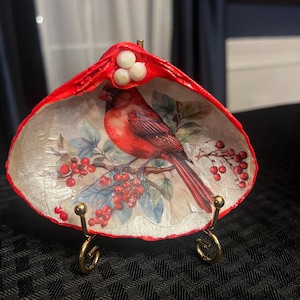 Può includere: Conchiglia decorativa dipinta di rosso all'esterno, con un'immagine dettagliata di un cardinale rosso appollaiato su un ramo con bacche rosse all'interno. La conchiglia è esposta su un piccolo supporto dorato.