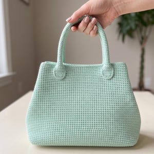 Peut inclure: Un sac à main vert menthe doté d'un corps rectangulaire structuré avec un motif de points au crochet dense et uniforme. Le sac comprend deux poignées supérieures arrondies et robustes, fixées au corps par des pattes ovales renforcées.