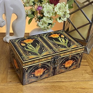 May include: A rectangular black storage box featuring a decorative floral pattern of orange tulips with green stems and leaves. The box has a hinged lid, a metallic latch closure on the front, and gold-toned trim accents along the edges and corners.