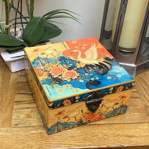 May include: A decorative wooden box with a hinged lid, featuring a Japanese-inspired design. The box is adorned with illustrations of cranes, flowers, and landscapes in shades of orange, blue, and yellow. The box is secured with a metal clasp.