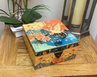 Caja cuadrada de madera decorada a mano en estilo decoupage. Presenta un exquisito diseño de garza japonesa en tonos melocotón, naranja y azul.