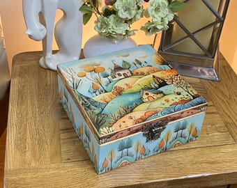 Caja cuadrada de madera decorada a mano en estilo decoupage. Presenta un paisaje caprichoso en tonos cálidos de azul, melocotón y verde.