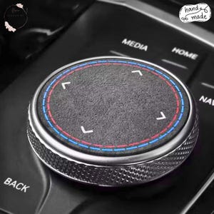 Può includere: Una copertura circolare per la manopola della console dell'auto con una superficie grigio scuro testurizzata e quattro frecce direzionali bianche. Il bordo ha una finitura metallica argentata zigrinata e un bordo circolare cucito rosso e blu.