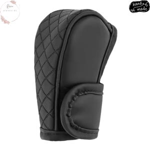 May include: Black shift knob cover with a quilted pattern. The cover is made of a smooth material and features a stitched design. The words "hand made" are in a black circle on the upper right.