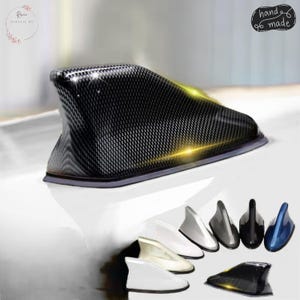 May include: A black carbon fiber car antenna in the shape of a shark fin. The image also shows several other car antennas in white, silver, gray, black, and blue. The words "hand made" are in the upper right corner.