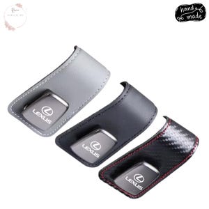 May include: Three leather key fob holders in grey, black, and carbon fibre. Each holder has a metal Lexus key fob inside. The grey holder has white stitching, the black has black stitching, and the carbon fibre has red stitching. The words "hand made" are in the top right corner.