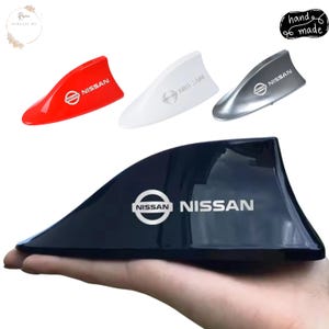 May include: Four shark fin car aerials in red, white, silver, and black. Each aerial features the Nissan logo and the word NISSAN printed in a metallic finish. The aerials have a smooth, aerodynamic shape designed for vehicle roof mounting.