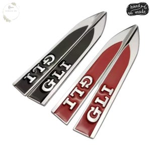 Puede incluir: Dos emblemas de coche, uno negro y otro rojo, cada uno con la palabra "GLI" en blanco. Los emblemas tienen un borde plateado y una forma elegante y alargada. La imagen está sobre un fondo blanco.