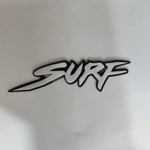May include: A white "SURF" graphic with a black outline. The word is in a stylized font, likely for a sticker or decal, and is centered on a white background. The design is simple and bold.