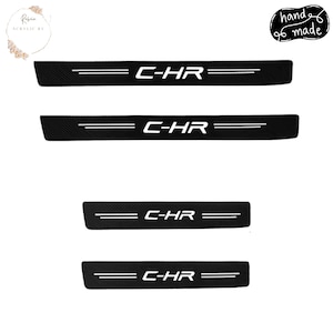 Pode incluir: Protetores de soleira de porta de carro pretos com o texto "C-HR" em branco. Os protetores têm uma textura de fibra de carbono e duas linhas brancas paralelas. As palavras "hand made" também são visíveis.