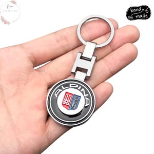 Pode incluir: Um porta-chaves de metal prateado com um emblema circular preto e prateado. O emblema apresenta a palavra "ALPINA" em letras brancas e um design vermelho e azul. O porta-chaves é segurado em uma mão. As palavras "hand made" estão no canto superior direito.