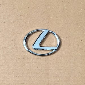 Puede incluir: Un emblema cromado de Lexus, con la "L" estilizada de la marca dentro de un óvalo. El emblema tiene un acabado metálico reflectante y está sobre un fondo neutro, de color beige. El diseño es limpio y moderno.
