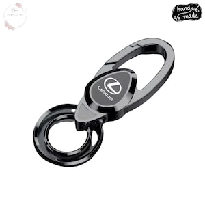 May include: A black and silver Lexus car key carabiner. The key ring has a spring-loaded clasp and a circular key holder. The Lexus logo is displayed on a black oval-shaped plate. The words "hand made" are written on the top right.