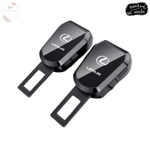 Puede incluir: Dos inserciones negras para hebillas de cinturón de seguridad de coche con el logotipo blanco de Lexus. Las inserciones son rectangulares con una parte superior curvada y una hebilla metálica. El texto "hand made" está en un círculo negro.