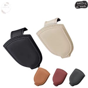 Op de afbeelding: Een verzameling van vijf schildvormige magnetische clips in zwart, crème, beige, bordeauxrood en donkergrijs. Elke clip heeft een glad, effen oppervlak met gestikte randen en een lus aan de bovenkant voor bevestiging.