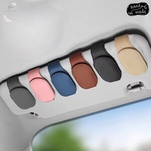 May include: Six oval-shaped car visor sunglass holders in various colours including dark grey, light pink, blue, brown, black, and beige. Each holder features a magnetic closure mechanism and is attached to a light grey vehicle sun visor.