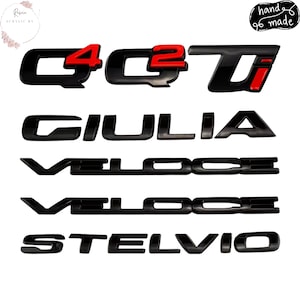以下が含まれることがあります： 「Q4」「Q2」「Giulia」「Veloce」「Stelvio」の文字が入った黒と赤の車のエンブレム。 「Q4」と「Q2」のエンブレムには赤いアクセントがあります。 画像には「hand made」の文字も含まれています。