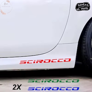 Puede incluir: Calcomanías "SCIROCCO" rojas, verdes y azules para un coche, expuestas en un vehículo blanco. La imagen también muestra una pegatina "hand made". Las calcomanías son probablemente para la personalización del coche.
