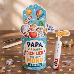 Puede incluir: Un recipiente cilíndrico de papel decorado con ilustraciones espaciales que incluyen un cohete, un sol y un planeta. El texto dice: PAPA, WIR HABEN DICH LIEB, BIS ZUM MOND & ZURÜCK. Una bandera dice BESTER PAPA. Una barra de chocolate separada tiene una estrella y el texto BIST UNERSETZLICH.