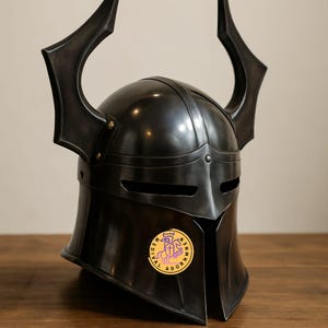 Könnte beinhalten: Ein dunkelgrauer mittelalterlicher Helm mit großen, gebogenen Hörnern. Der Helm hat eine abgerundete Oberseite, Augenschlitze und einen Nackenschutz. Ein goldfarbenes Emblem ist an der Seite angebracht. Der Helm ist aus Metall und hat eine polierte Oberfläche.