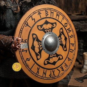 May include: A circular, tan shield featuring black runic inscriptions and a central black design. It has a silver metal boss in the middle. The shield is held by a person wearing a fur-trimmed garment.