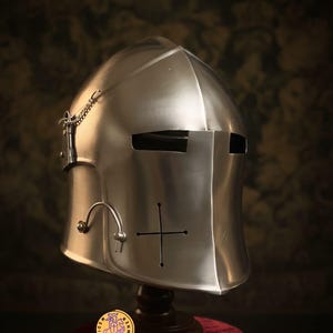 May include: A shiny silver medieval helmet with a cross design and eye slits. The helmet is displayed on a wooden stand with a red velvet cloth. A small round emblem is at the base.