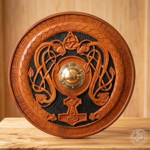 May include: A circular wooden shield featuring intricate carvings of two intertwined serpents and a central hammer symbol. The design includes a polished brass dome at the center, surrounded by a border of etched runic characters and braided patterns.