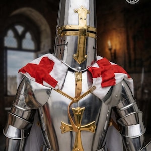 A polished steel suit of armor featuring a cylindrical great helm with a brass cross, a breastplate adorned with a large brass cross emblem, and a white mantle draped over the shoulders with red cross patches on each side.